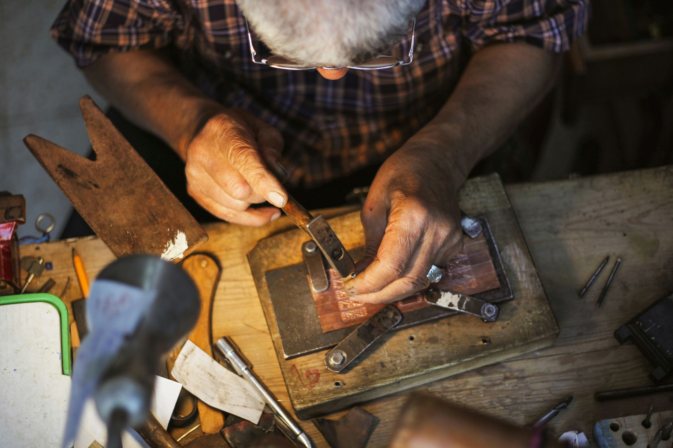 Close-up of an artisan's hands engraving a copper plate with a hammer and chisel at a wooden workbench